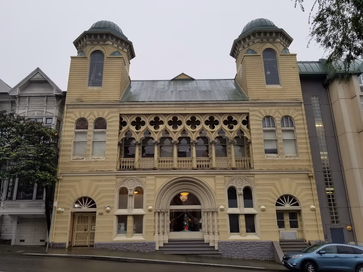 Front exterior view of a historic, ornate building with two towers, arched windows, and decorative architectural details. The building is painted yellow with a greenish roof and is situated on a street with a parked car visible on the right.