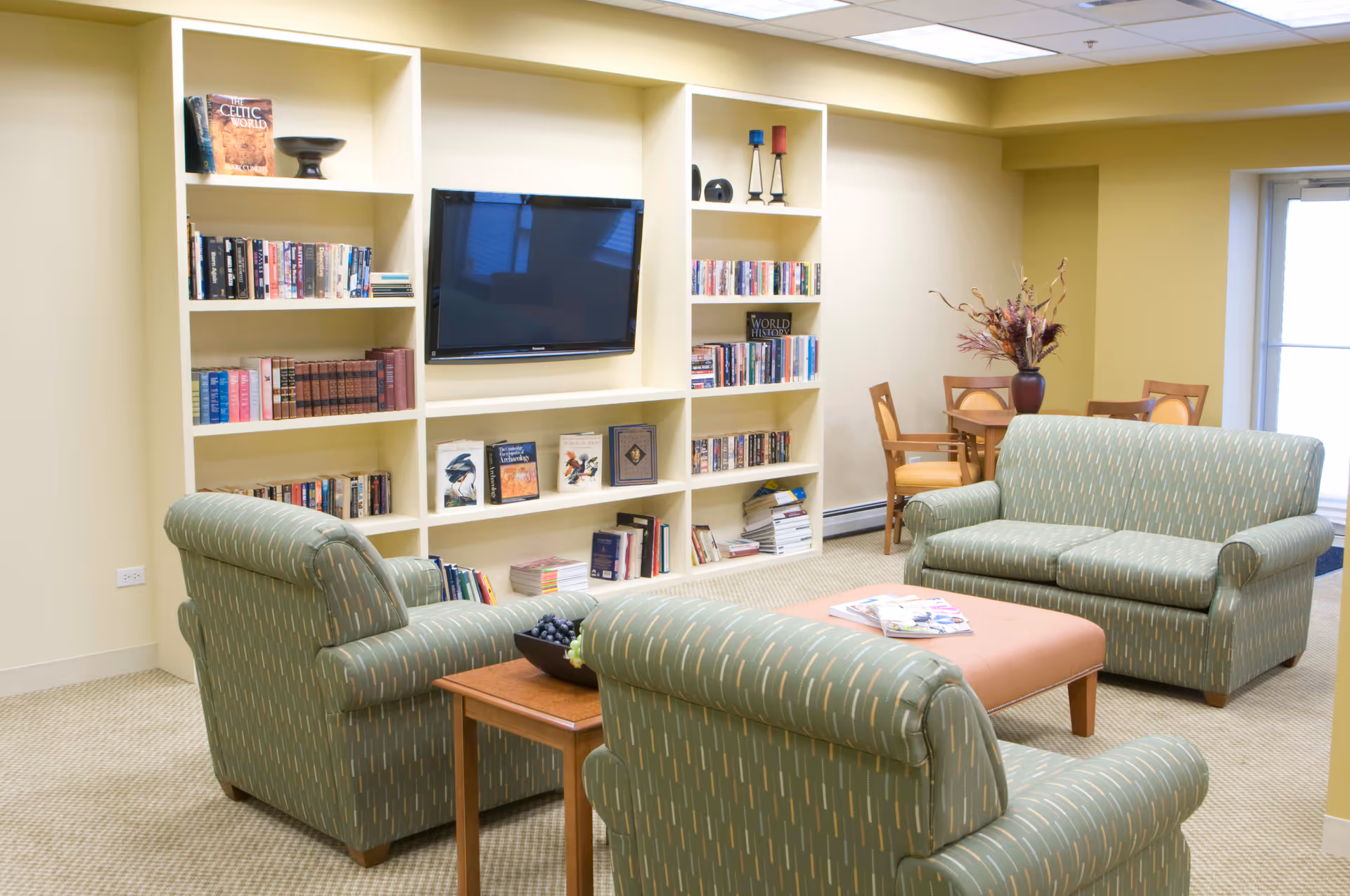 A cozy living room area in a senior living facility with three green patterned armchairs and a matching loveseat arranged around a rectangular ottoman. Behind the seating is a built-in bookshelf filled with books and a flat-screen TV mounted in the center. In the background, there is a small round table with four wooden chairs and a vase with flowers. The room has beige walls and carpeted flooring.