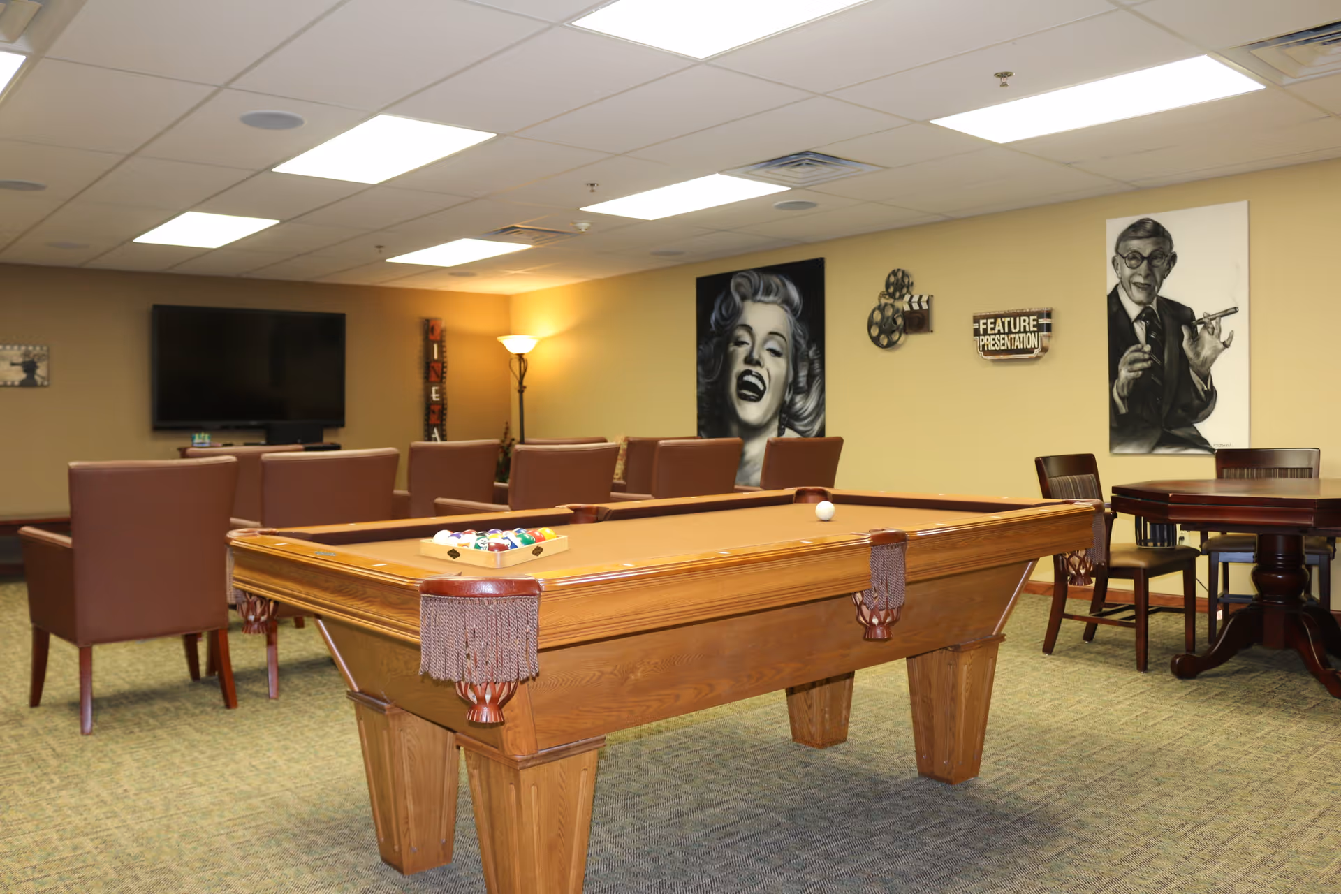 A recreational room with a pool table in the center, surrounded by brown chairs and a table. The walls are decorated with black and white portraits and a sign that reads 'Feature Presentation'. A large flat-screen TV is mounted on the far wall, and the room is well-lit with ceiling lights and a floor lamp.
