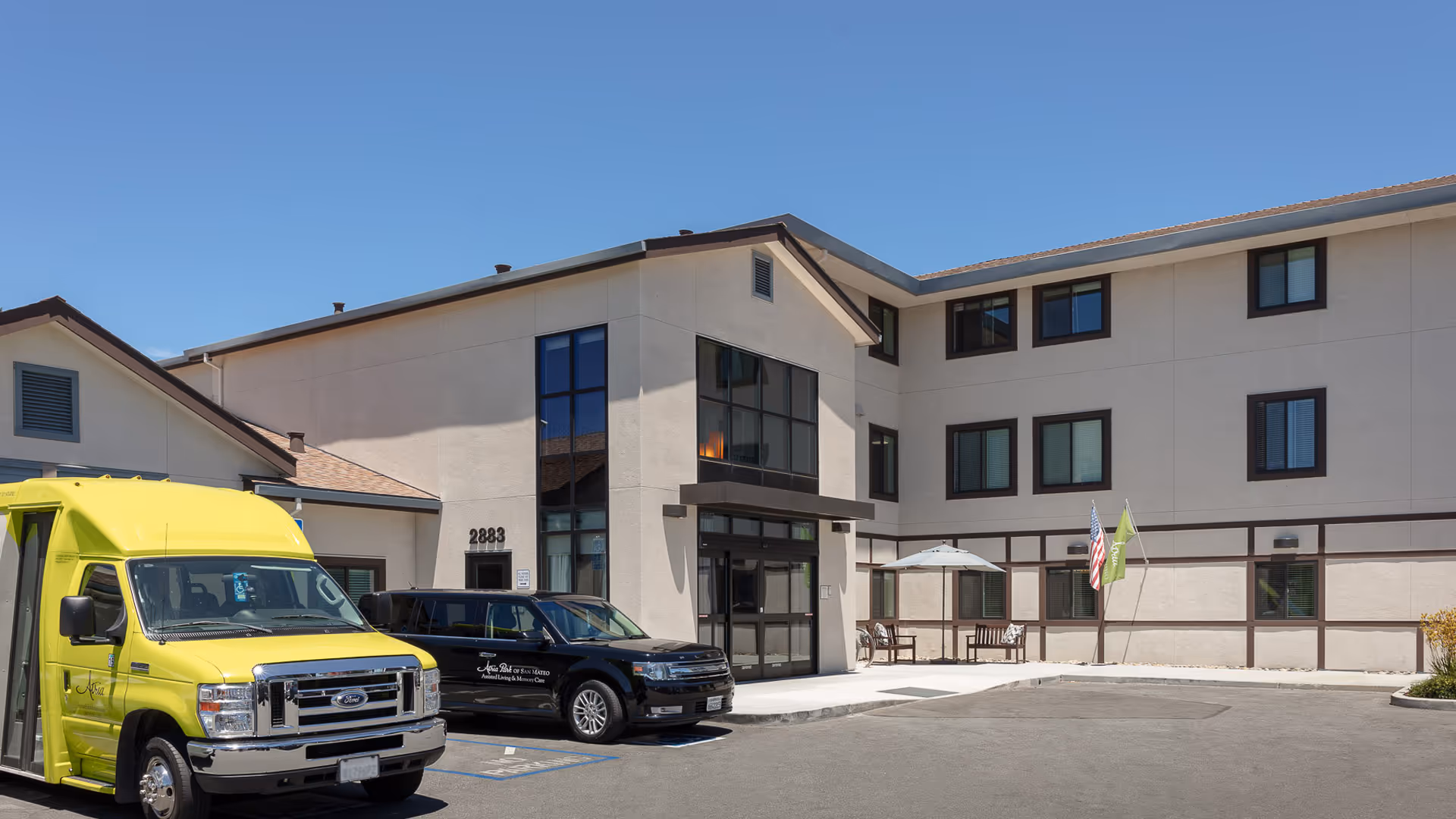 Exterior view of Atria Park of San Mateo building with a yellow shuttle bus and a black vehicle parked in front. The building has three stories with multiple windows and a small seating area with an umbrella near the entrance. Two flags, one American and one green, are displayed near the seating area.