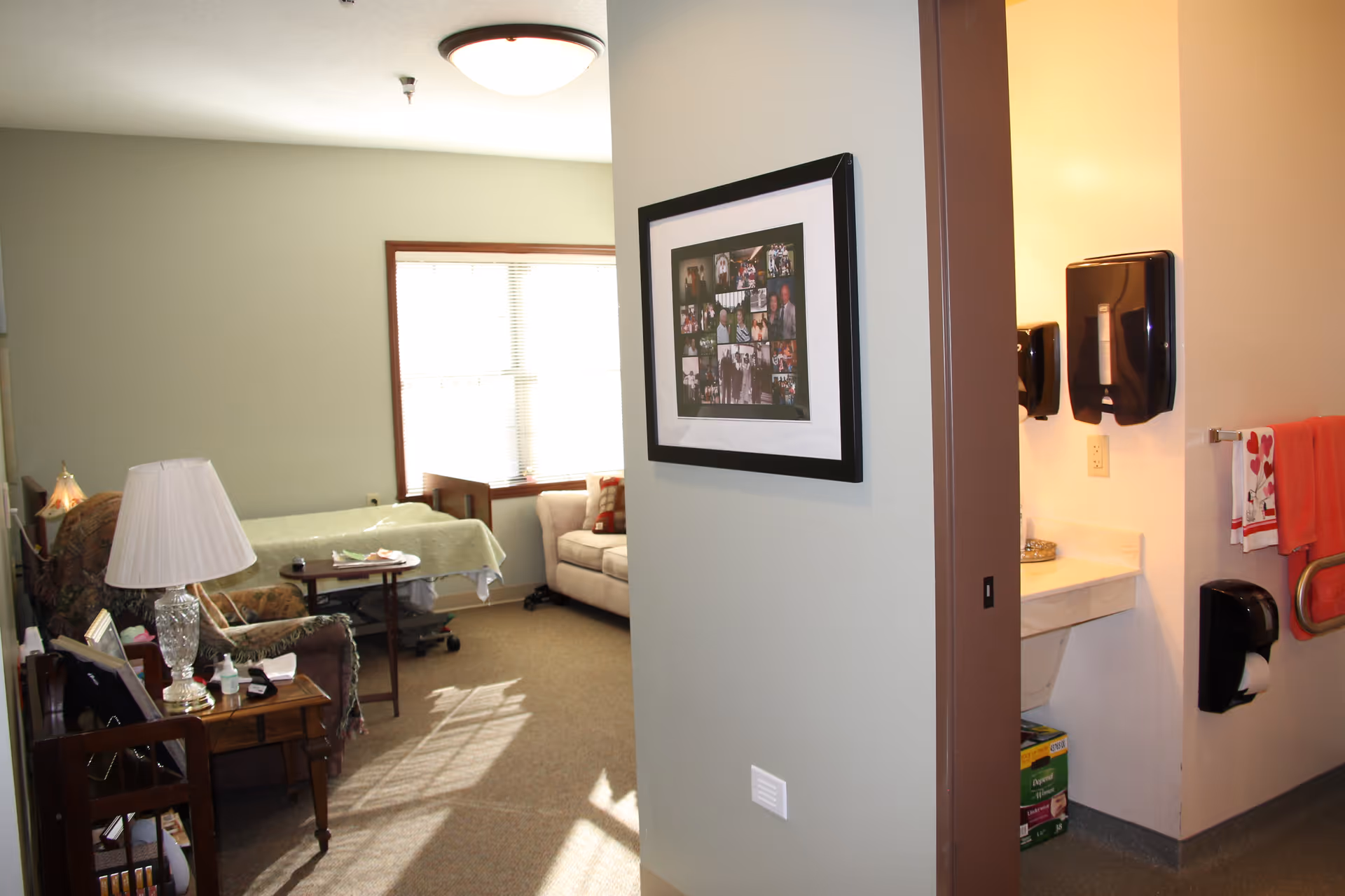 Interior view of a senior living resident room with a bed, seating area, lamp, and an adjacent bathroom sink visible through a doorway.