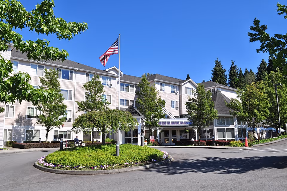 Exterior view of Hillside Assisted Living facility on a clear sunny day, showing a three-story building with multiple windows, surrounded by green trees and landscaping. An American flag is flying on a flagpole in front of the building, and there is a circular driveway with a landscaped island in the center.