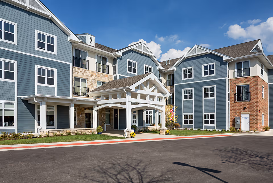 Exterior view of a modern senior living facility building with blue and beige siding, multiple windows, balconies, and a covered entrance with white pillars. The sky is clear with a few clouds, and there is a paved driveway in front.