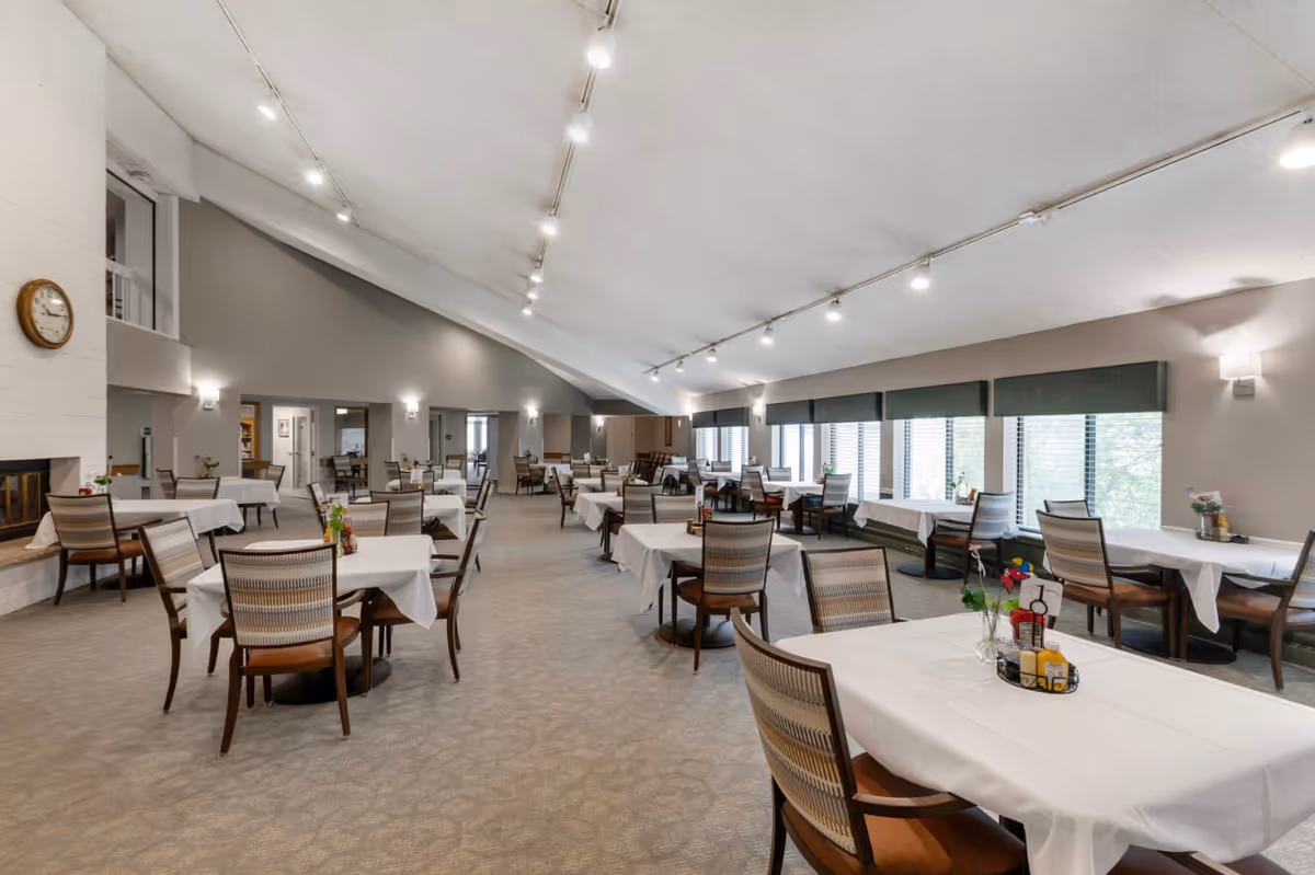 A spacious dining room in a senior living facility with multiple tables covered in white tablecloths and surrounded by chairs. The room features large windows with blinds, a high slanted ceiling with track lighting, and a wall clock above a fireplace on the left side.