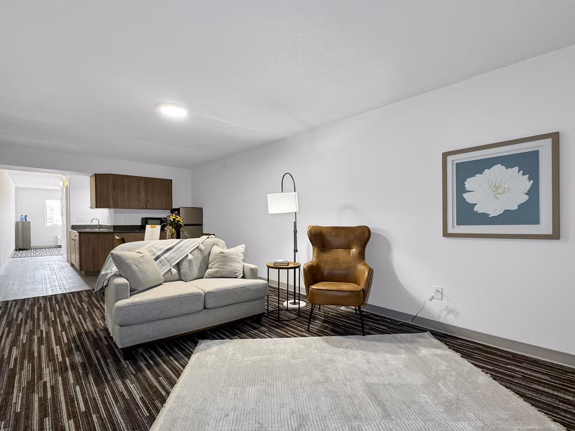 Modern living room with a gray sofa, brown leather armchair, floor lamp, framed floral artwork on the wall, and a kitchenette visible in the background.