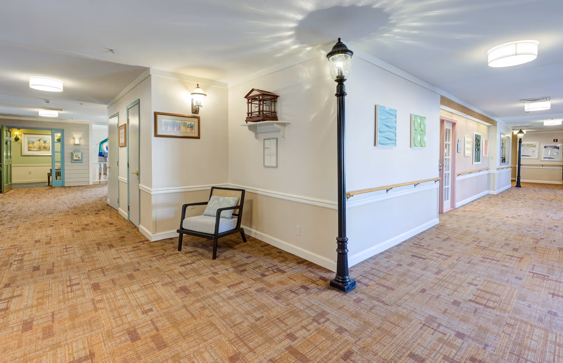 A well-lit hallway in a senior living facility with beige walls and patterned carpet flooring. The hallway features a black streetlamp-style light fixture, a single cushioned chair, framed artwork on the walls, and handrails along the corridor. Doors and decorative elements are visible along the hallway, creating a welcoming atmosphere.