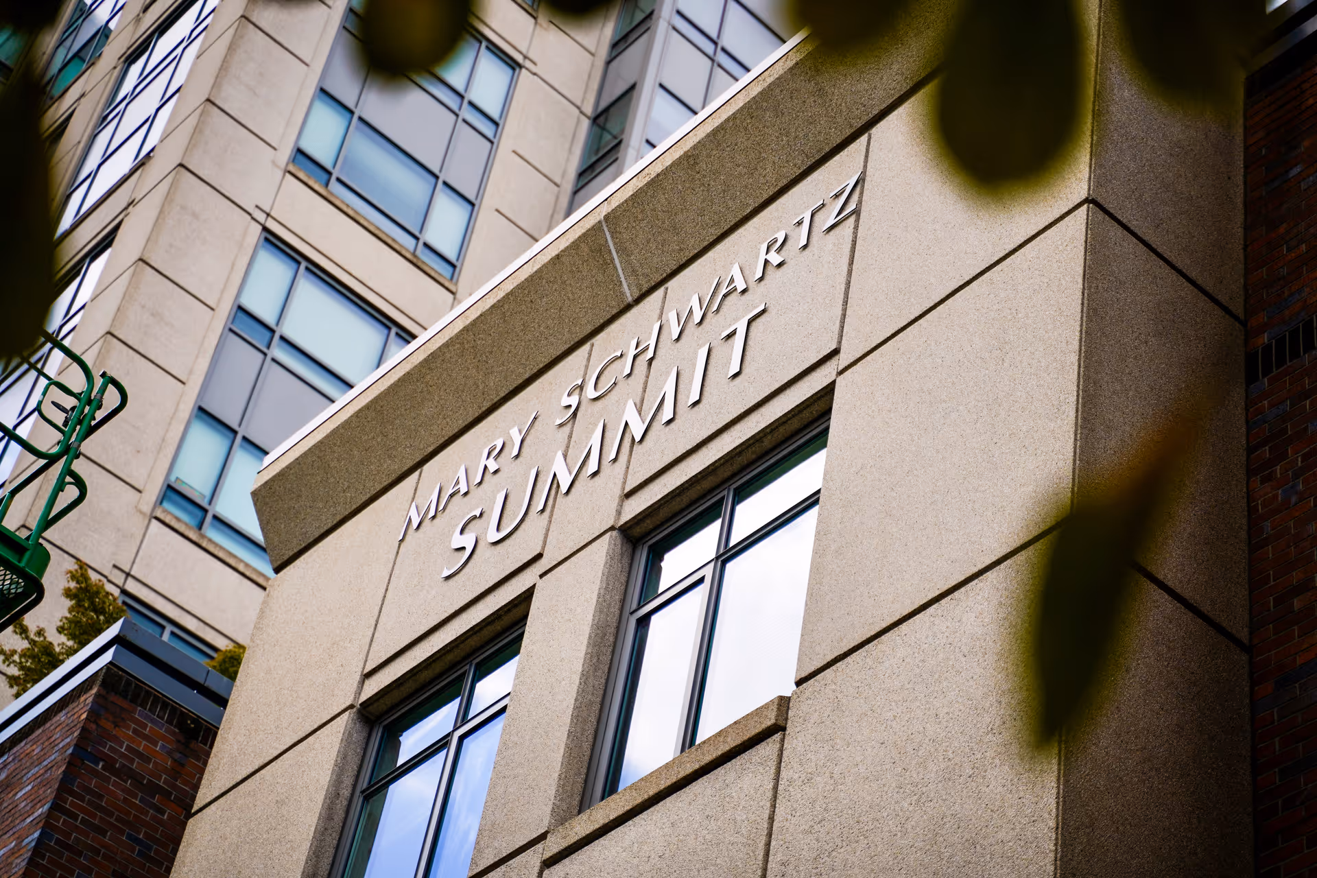 Exterior view of a building with the name 'Mary Schwartz Summit' displayed prominently above windows, with parts of other buildings and some blurred leaves in the foreground.