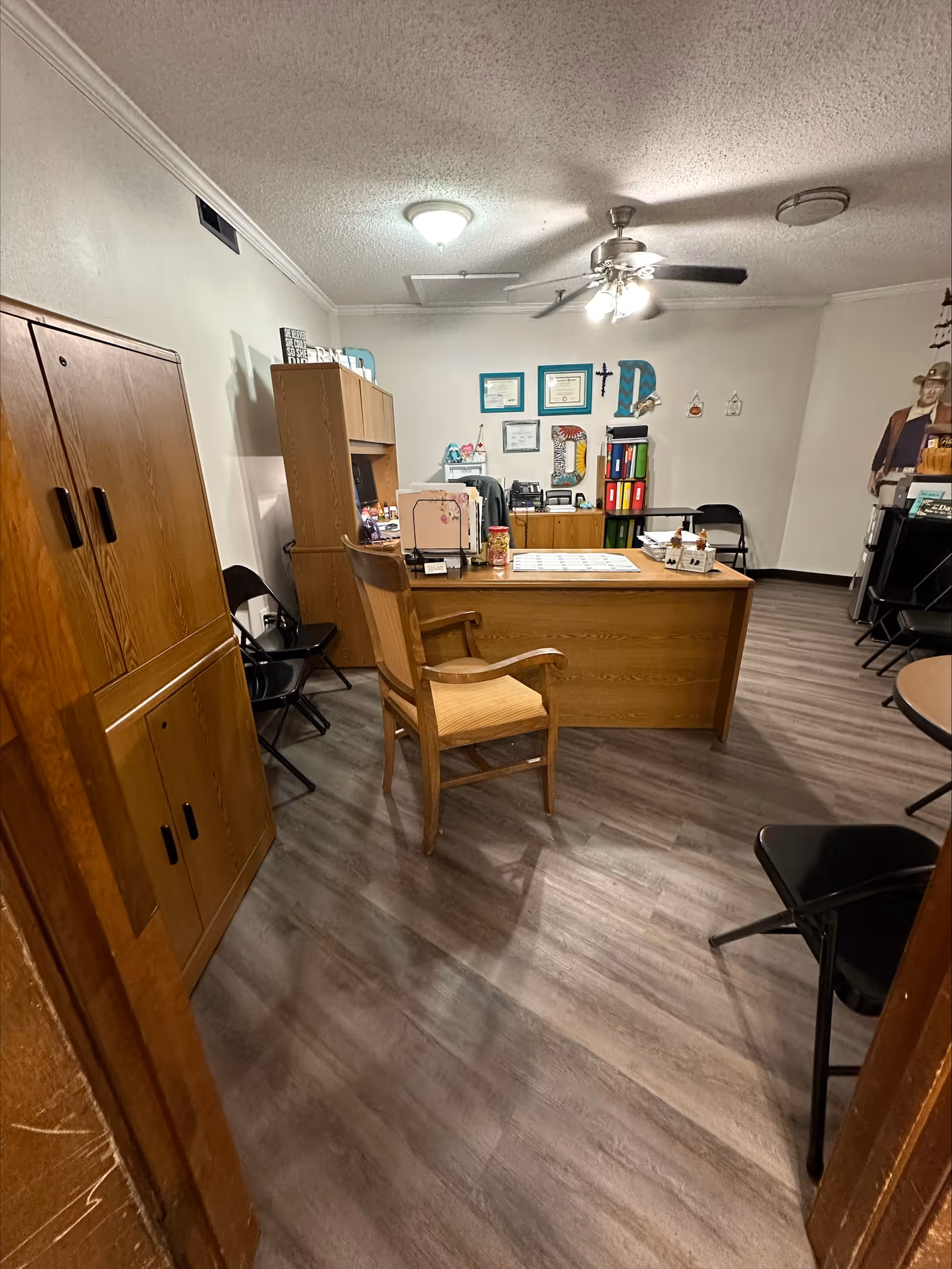 An office room with a wooden desk and chair in the center, surrounded by black folding chairs. There is a wooden cabinet on the left and a wooden shelving unit behind the desk with various office supplies and decorations. The walls have framed certificates and colorful letters, and the ceiling has a fan with lights. The floor is covered with wood-patterned vinyl flooring.