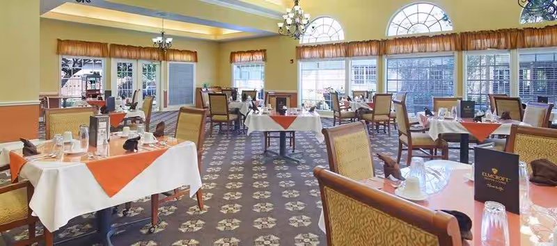 A spacious dining room with multiple tables covered in white tablecloths and orange table runners. Each table is set with glasses, cups, and menus. The room features large arched windows with orange valances, chandeliers, and patterned carpet flooring.