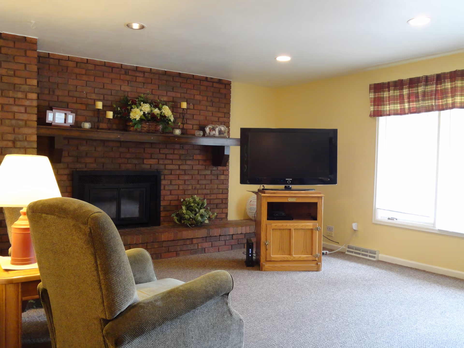 A cozy living room with a brick fireplace decorated with candles and a flower arrangement on the mantel. There is a comfortable armchair facing a wooden TV stand with a flat-screen television. A window with a plaid valance allows natural light into the room, which has yellow walls and carpeted flooring.