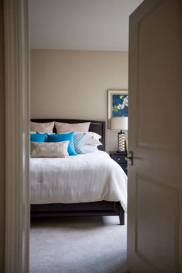 Partially open door revealing a neatly made bedroom with a bed, colorful pillows, nightstand, and lamp.