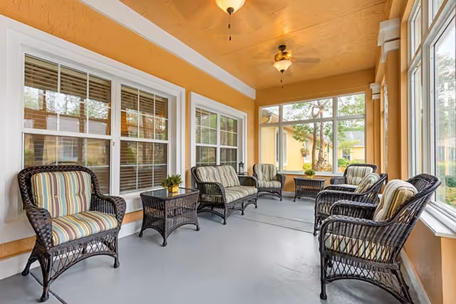 A bright enclosed patio area with wicker chairs and sofas featuring striped cushions arranged around small wicker tables. The walls are painted a warm yellow-orange color, and large windows let in natural light, showing greenery outside.