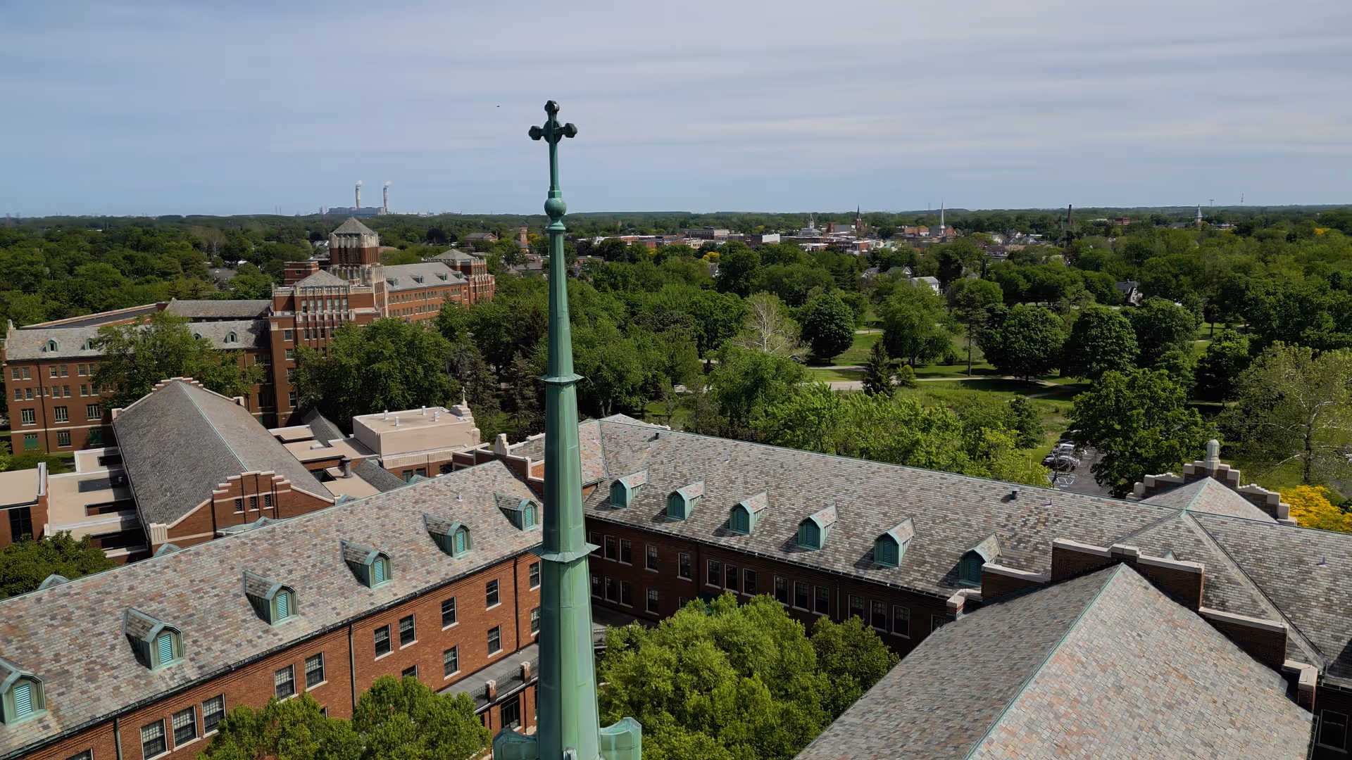 Aerial view of a large brick building complex with a tall green steeple topped with a cross, surrounded by trees and greenery under a partly cloudy sky.