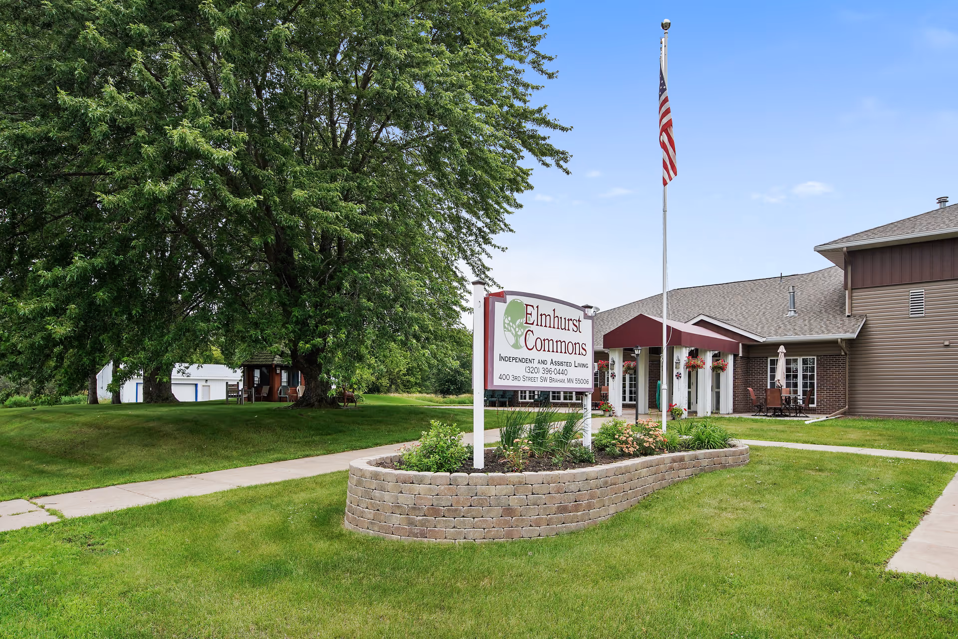Entrance to Elmhurst Commons senior living building with a landscaped sign, American flagpole, and large tree on a grassy lawn.