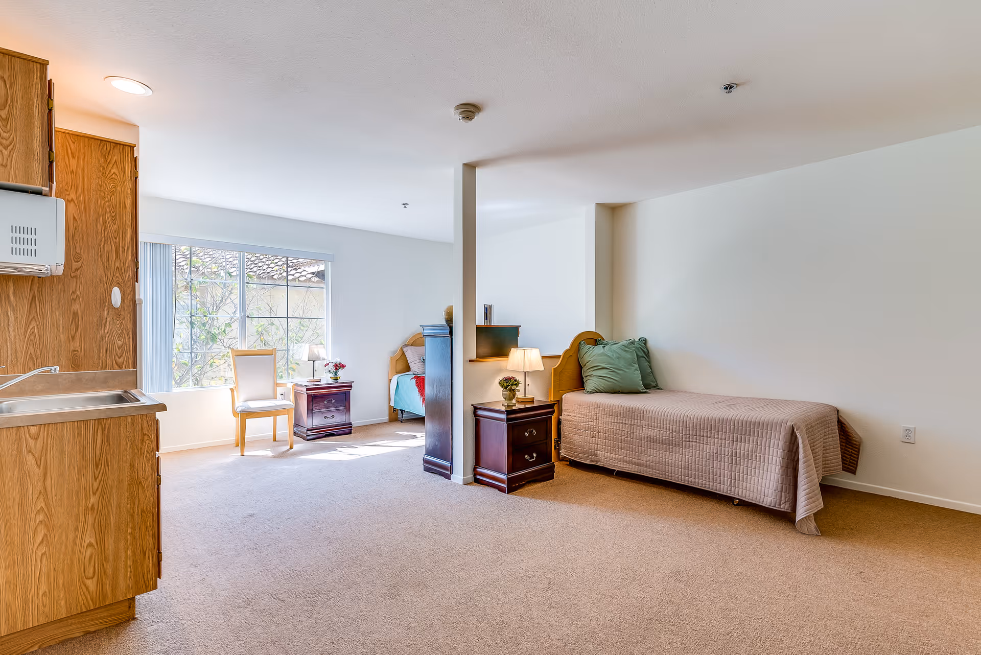 Studio-style assisted living room with a single bed, nightstands, a chair by a large window, and a small kitchenette.