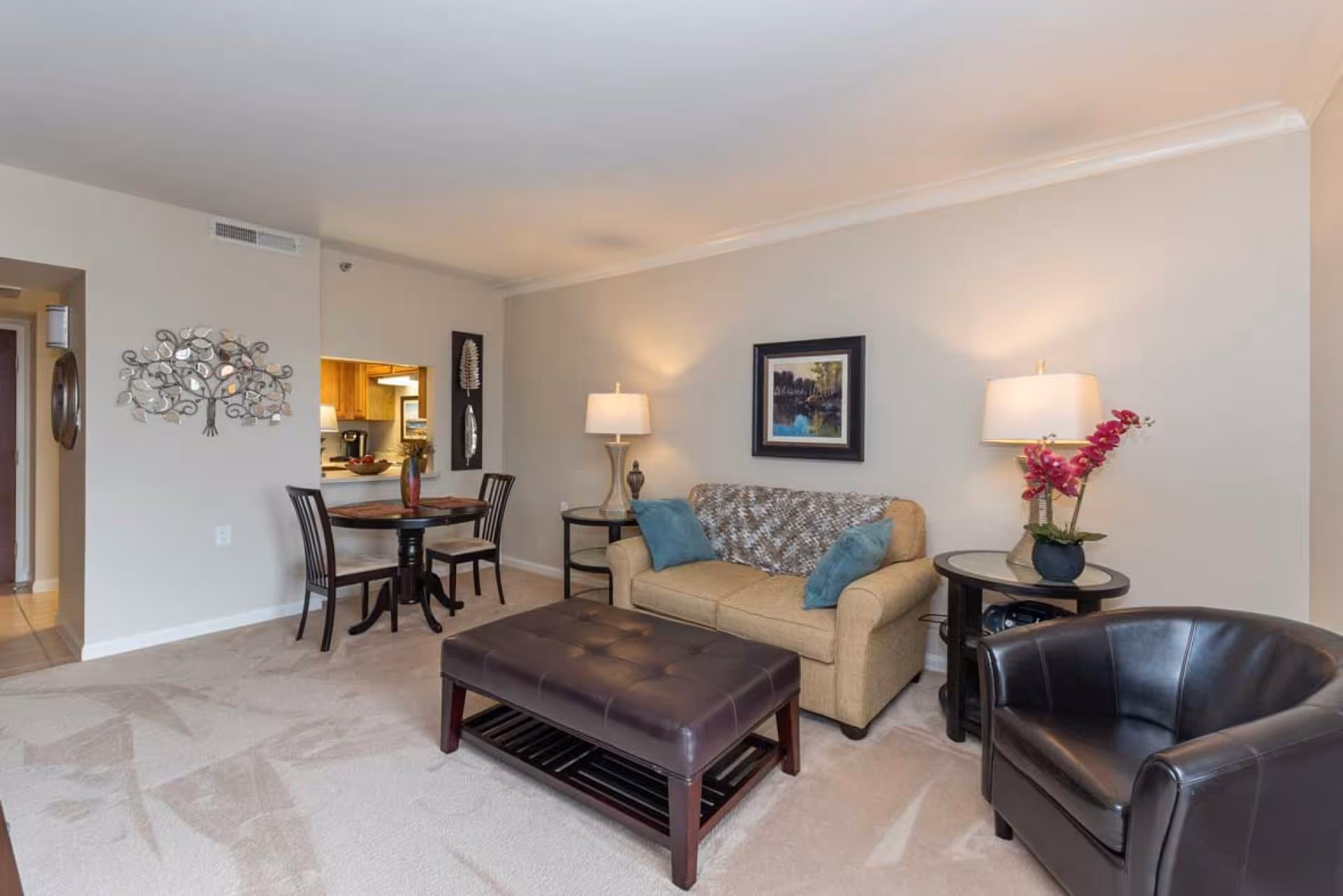 Well-lit living room with a sofa, ottoman, leather armchair, side tables and lamps, and a small dining table in the background.