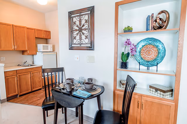 A small dining area with a dark wooden table set for two with dishes, cups, and napkins. Two black wooden chairs are placed around the table. To the left is a kitchenette with wooden cabinets, a small refrigerator, microwave, and sink. On the right, there is a built-in wooden shelving unit displaying decorative items including a blue patterned plate, a purple orchid, books, and small boxes. A decorative metal wall art piece hangs on the wall above the table.