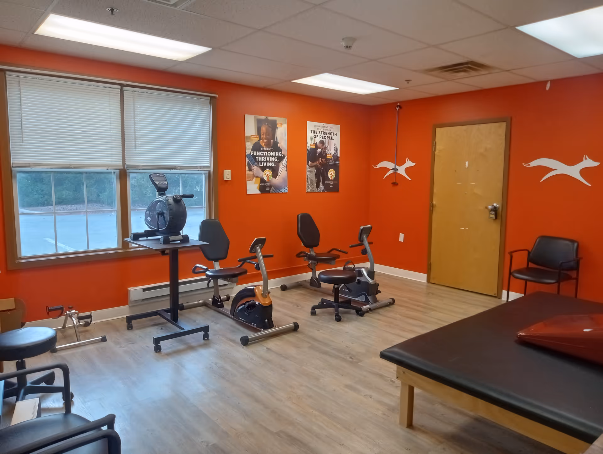 A small exercise room with orange walls and wood flooring. The room contains two stationary exercise bikes, a rowing machine, a black padded exercise table, and several chairs. Two motivational posters are on the wall, and there are white fox-like decals on the walls near a wooden door. A large window with closed blinds lets in natural light.