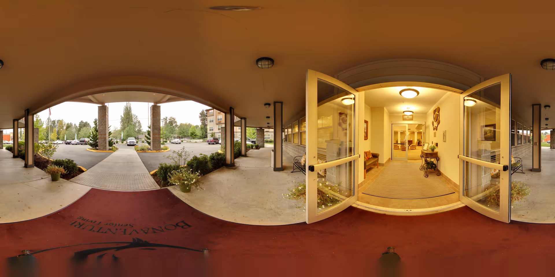 Entrance of North Creek by Bonaventure senior living facility showing open glass double doors leading into a well-lit lobby area with seating and decorative items. Outside, there is a covered walkway with columns, potted plants, and a parking lot with cars and trees in the background.