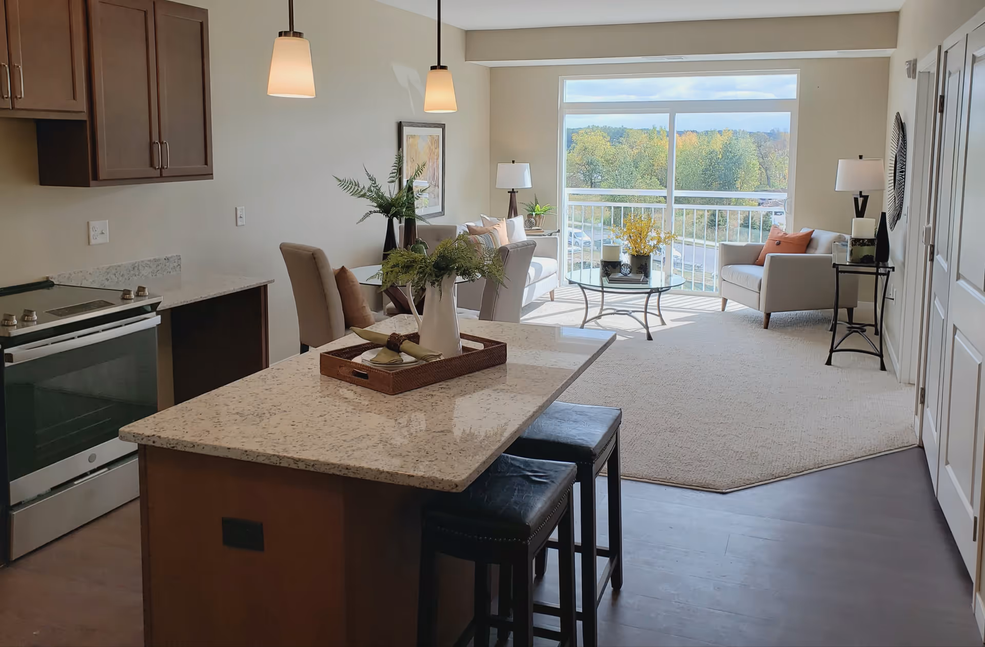 A bright and modern living space featuring a kitchen area with a granite countertop island and two black stools. The kitchen has wooden cabinets and a stainless steel stove. Beyond the kitchen is a dining area with a small table and chairs, and a living room with two armchairs, a glass coffee table, and large windows offering a view of trees and a parking lot outside. The room is decorated with plants, lamps, and wall art.