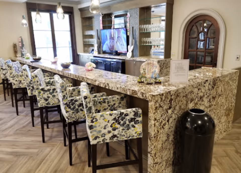 Interior view of a senior living facility's common area featuring a long granite countertop bar with floral upholstered bar stools. Behind the bar is a TV mounted on the wall with glass shelves holding glassware. Pendant lights hang from the ceiling, and there is a decorative arched wooden door on the right side.