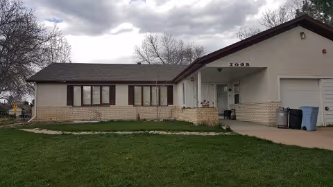 Single-story residential building with beige brick and light-colored siding, a covered front porch, and a garage. The lawn in front is green and well-maintained, with a cloudy sky overhead.