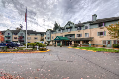 Exterior view of a three-story senior living facility building with a green awning over the main entrance labeled 'Brookdale Courtyard Puyallup.' There is a circular driveway with a flagpole and some landscaping in front, and several parked cars are visible on the left side. The sky is cloudy.