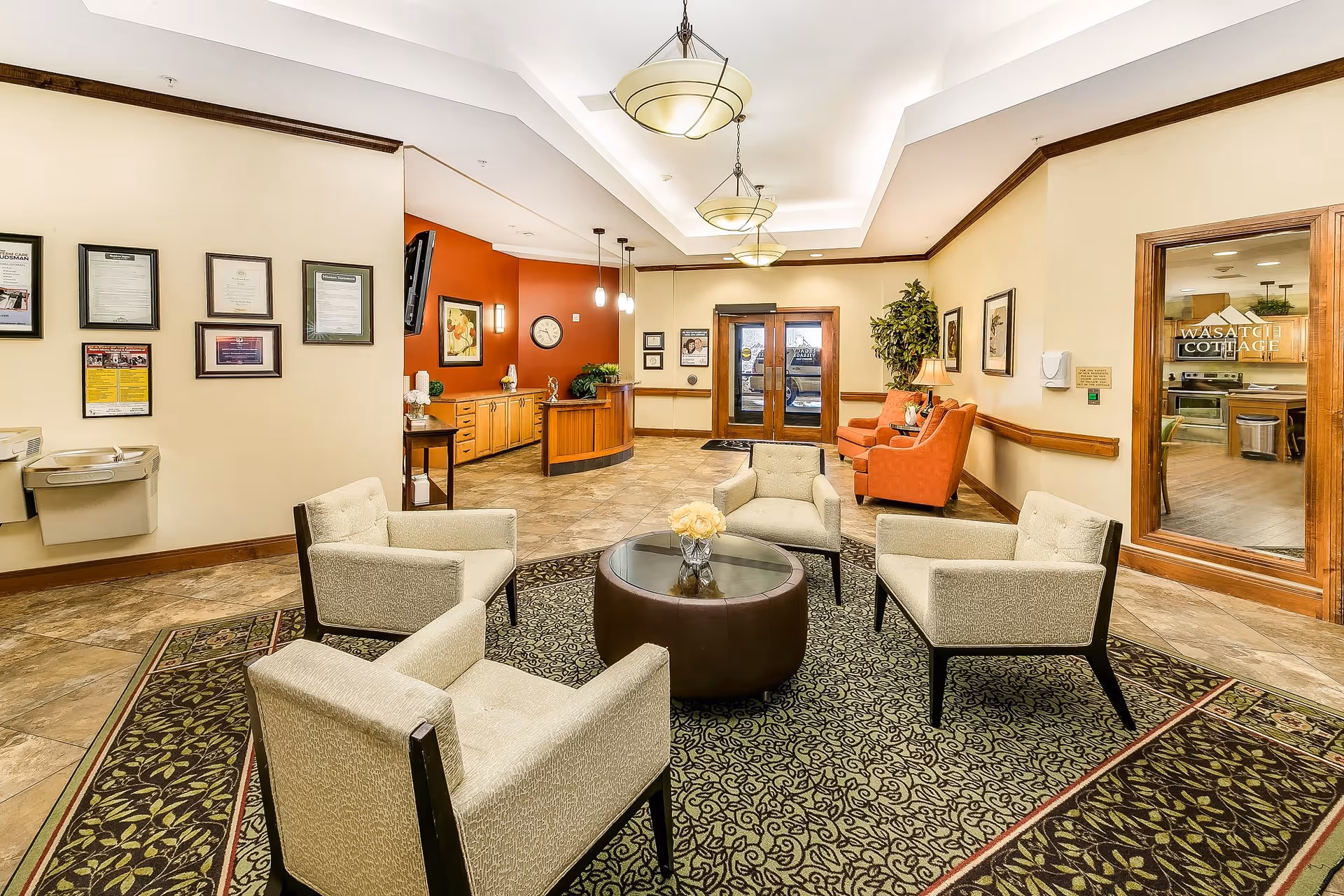 A bright and welcoming lobby area with four beige armchairs arranged around a round glass-top coffee table on a patterned rug. The walls are painted cream with a red accent wall behind a wooden reception desk. There are framed certificates and artwork on the walls, a water fountain, and double glass doors leading outside. To the right, a glass door labeled 'Wasatch Cottage' reveals a kitchen area.