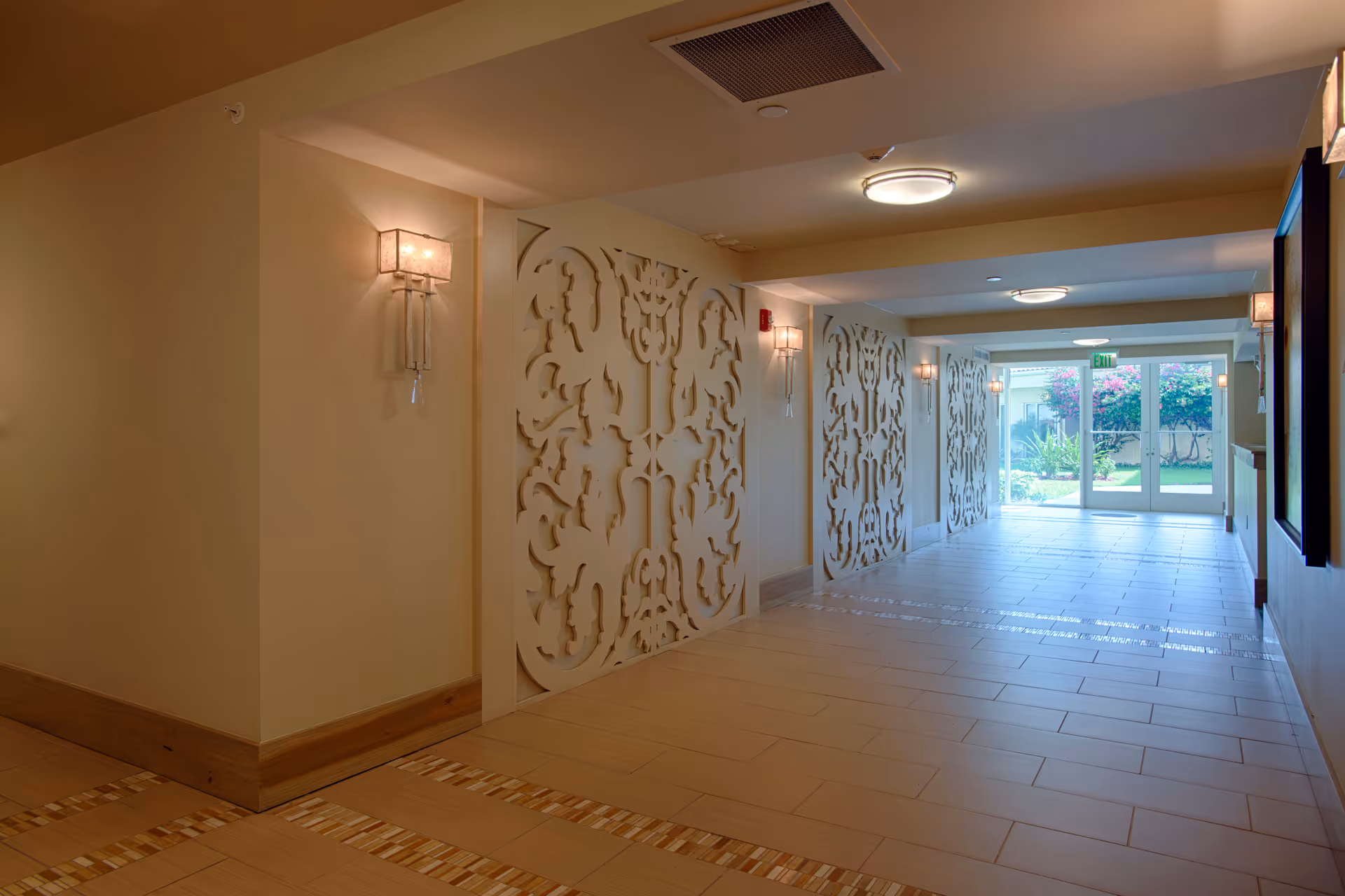 A well-lit hallway with decorative wall panels featuring intricate cut-out designs. The floor is tiled with light-colored tiles and has a patterned border. Wall sconces with soft lighting are mounted along the walls. At the end of the hallway, there are glass double doors leading to an outdoor garden area with greenery and flowering bushes.