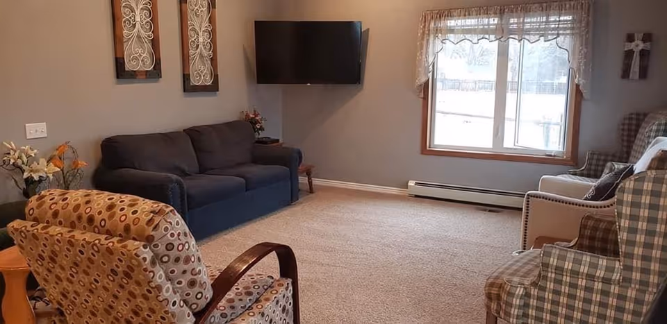 A cozy living room with a dark blue couch, two patterned armchairs, and a white armchair. A flat-screen TV is mounted on the wall above a small side table with flowers. There is a large window with sheer curtains letting in natural light. The walls are painted light gray and the floor is carpeted.