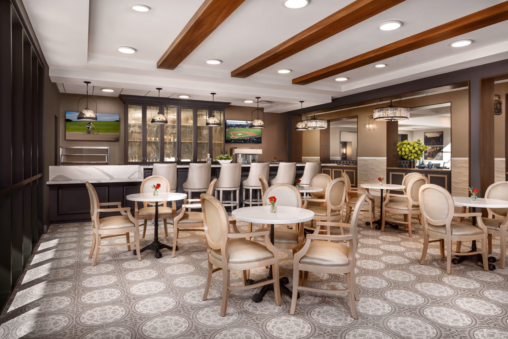 A bright and elegant dining area with round tables and beige upholstered chairs. Each table has a small vase with a red flower. In the background, there is a bar with high chairs, glass cabinets, and two wall-mounted televisions showing a cyclist and a baseball game. The ceiling features wooden beams and recessed lighting, and the floor has patterned tiles.
