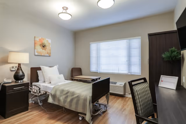 A clean and well-lit bedroom in a memory care community featuring a single hospital-style bed with white and beige bedding, a bedside table with a lamp and phone, a brown chair, a wooden desk with a chair, a flat-screen TV mounted on the wall, a window with blinds, and a wardrobe.