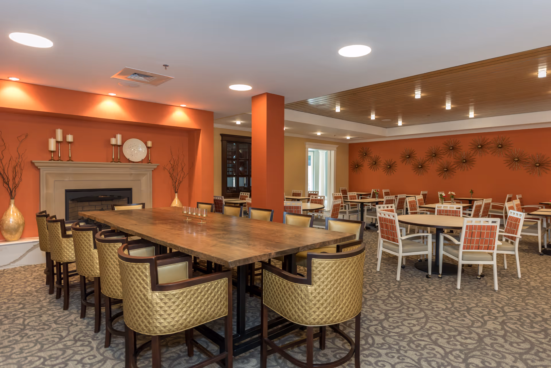 A spacious dining room with a large wooden table surrounded by cushioned chairs in the foreground. The room features an orange accent wall with a fireplace decorated with candles and vases. Additional round tables with chairs are arranged in the background, and the ceiling has recessed lighting and a wooden panel section.