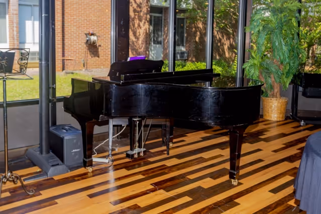 A black grand piano placed on a polished wooden floor inside a room with large glass windows showing an outdoor view with greenery and a brick building. There is a potted plant to the right of the piano and a metal stand to the left.