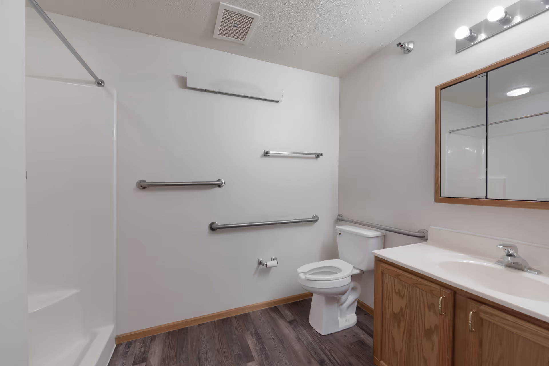 A clean bathroom with a white toilet, a wooden vanity with a sink, a large mirror above the sink, and a walk-in shower with a grab bar. The walls are white, and there are multiple stainless steel grab bars installed for accessibility. The floor has dark wood-style flooring.
