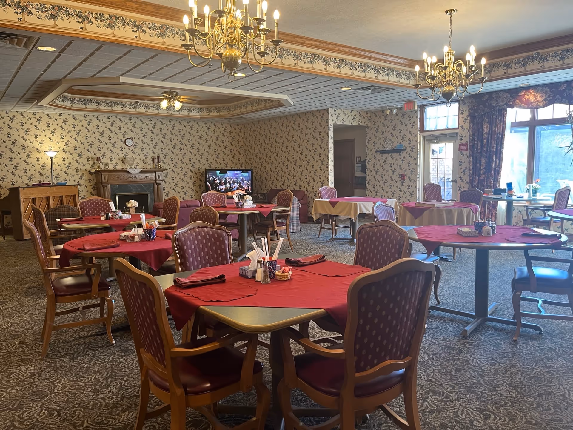 A dining room in a senior living facility with multiple tables covered in red and beige tablecloths. Each table is surrounded by wooden chairs with purple cushioned seats and backs. The room features floral wallpaper, a carpeted floor, chandeliers, and a television near a fireplace. Large windows with floral curtains allow natural light to enter the space.