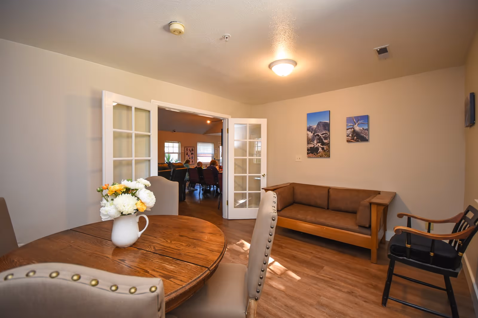 A cozy interior room with a round wooden table adorned with a white pitcher holding white and yellow flowers. Surrounding the table are beige upholstered chairs with nailhead trim. Against the wall is a wooden framed brown cushioned bench and a black wooden chair with armrests. Two landscape photos hang on the beige wall. French doors open to another room where several people are seated.