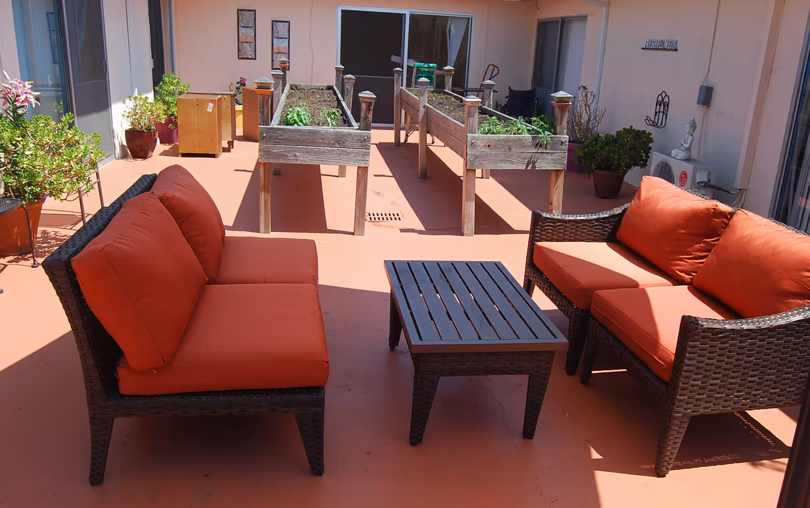 Outdoor patio area with two wicker sofas with orange cushions facing each other across a black slatted coffee table. Behind the seating area are two raised garden beds with plants. Various potted plants and decorative items are placed around the patio.