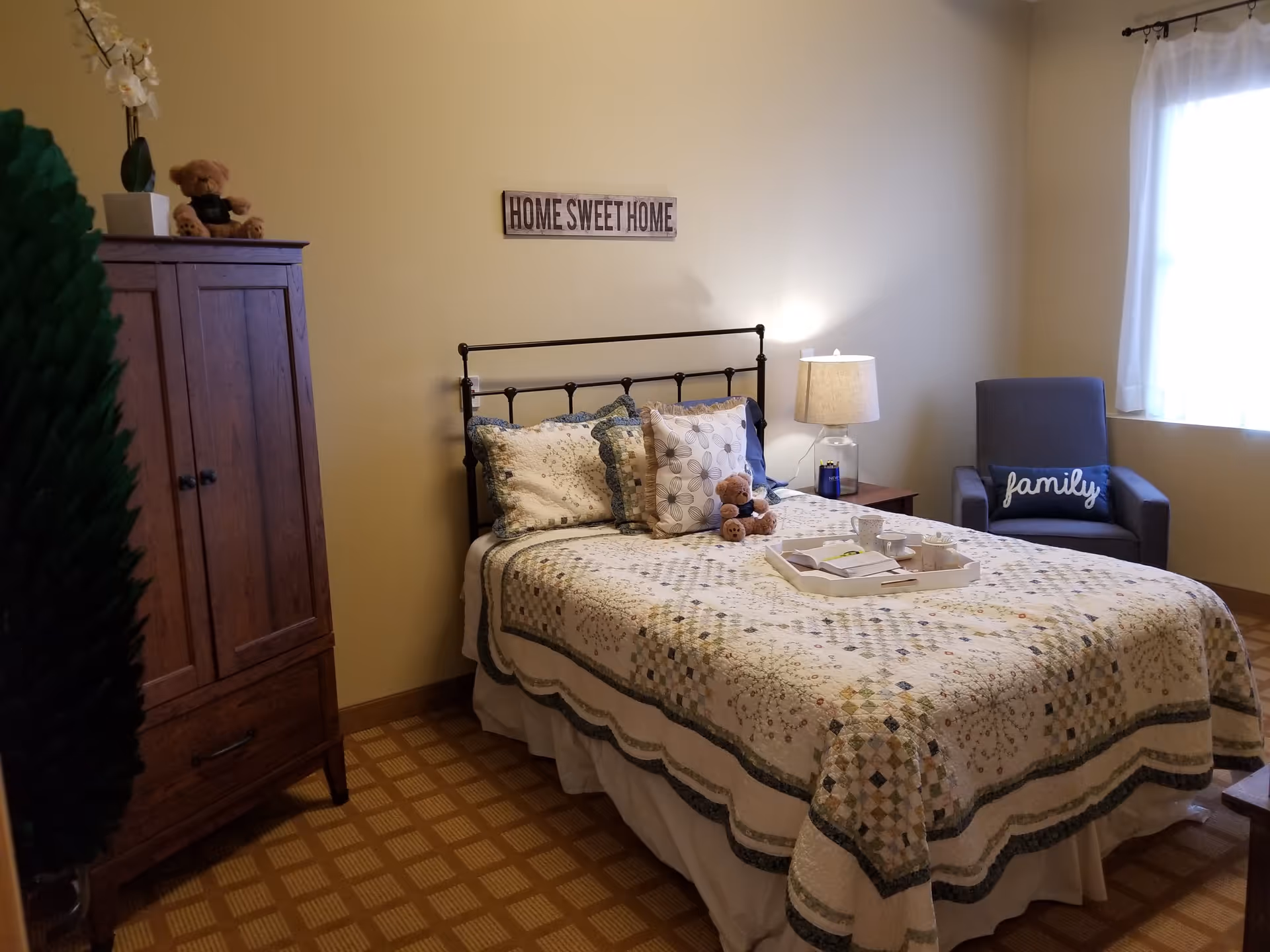 A cozy bedroom with a bed covered in a patterned quilt and multiple pillows, including a teddy bear. There is a wooden wardrobe on the left, a bedside table with a lamp, and a gray armchair with a pillow that says 'family' near a window with white curtains. A 'HOME SWEET HOME' sign is mounted on the wall above the bed.