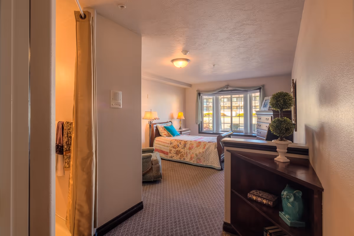 Furnished senior living bedroom with a bed, nightstand lamps, armchair and a large bay window.
