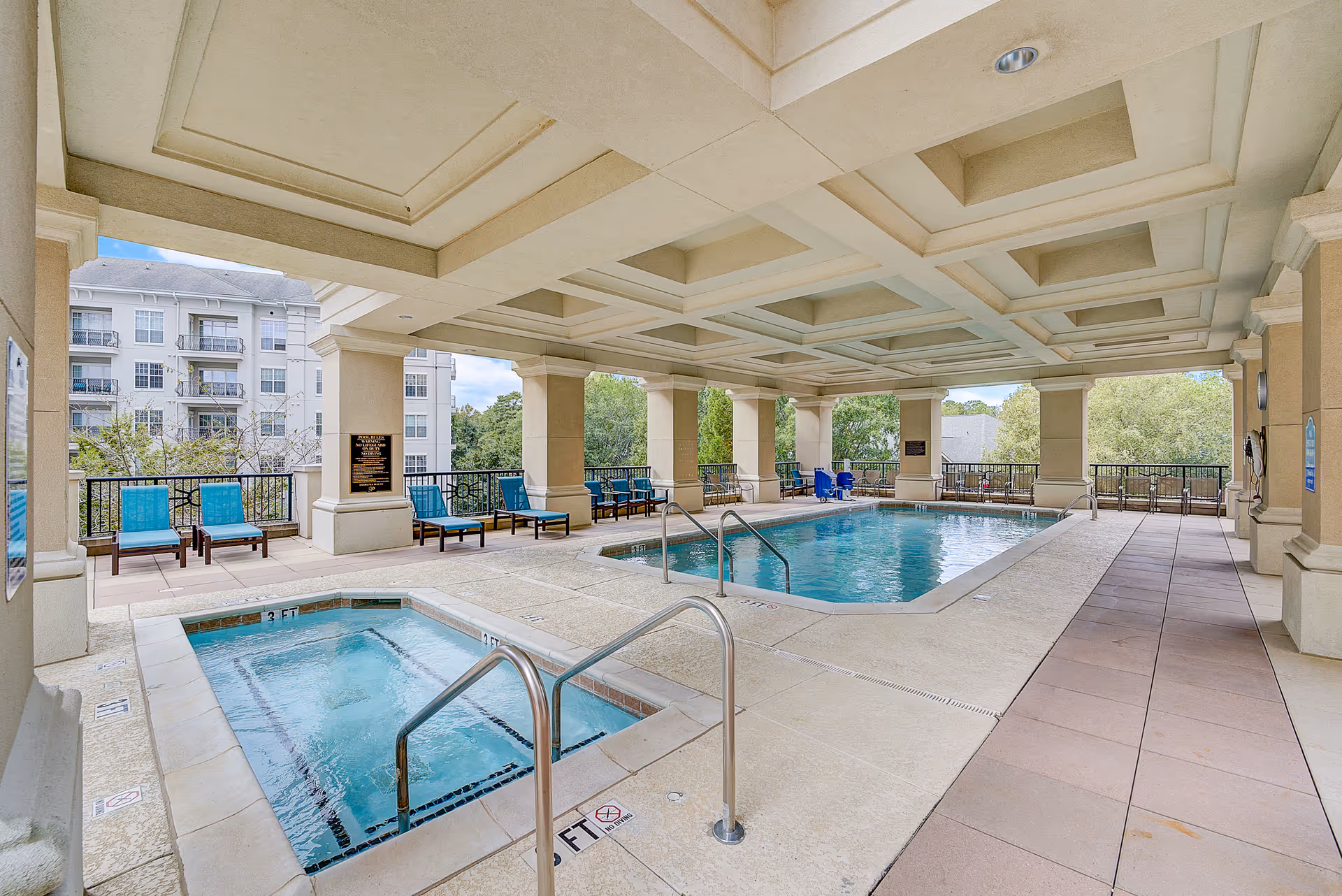 Covered pool and hot tub area with lounge chairs under a coffered ceiling overlooking nearby buildings and trees.