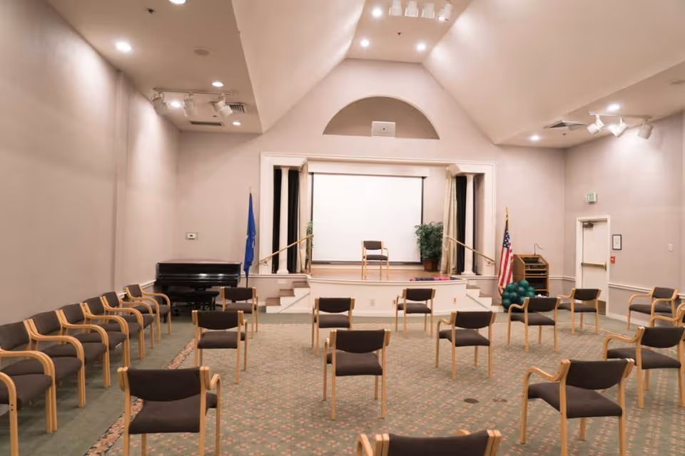A large meeting or activity room with multiple wooden chairs arranged in rows facing a small stage with a single chair. The room has a piano on the left side, an American flag on the right, and a projection screen on the stage. The walls are light-colored, and the ceiling has recessed lighting and spotlights.
