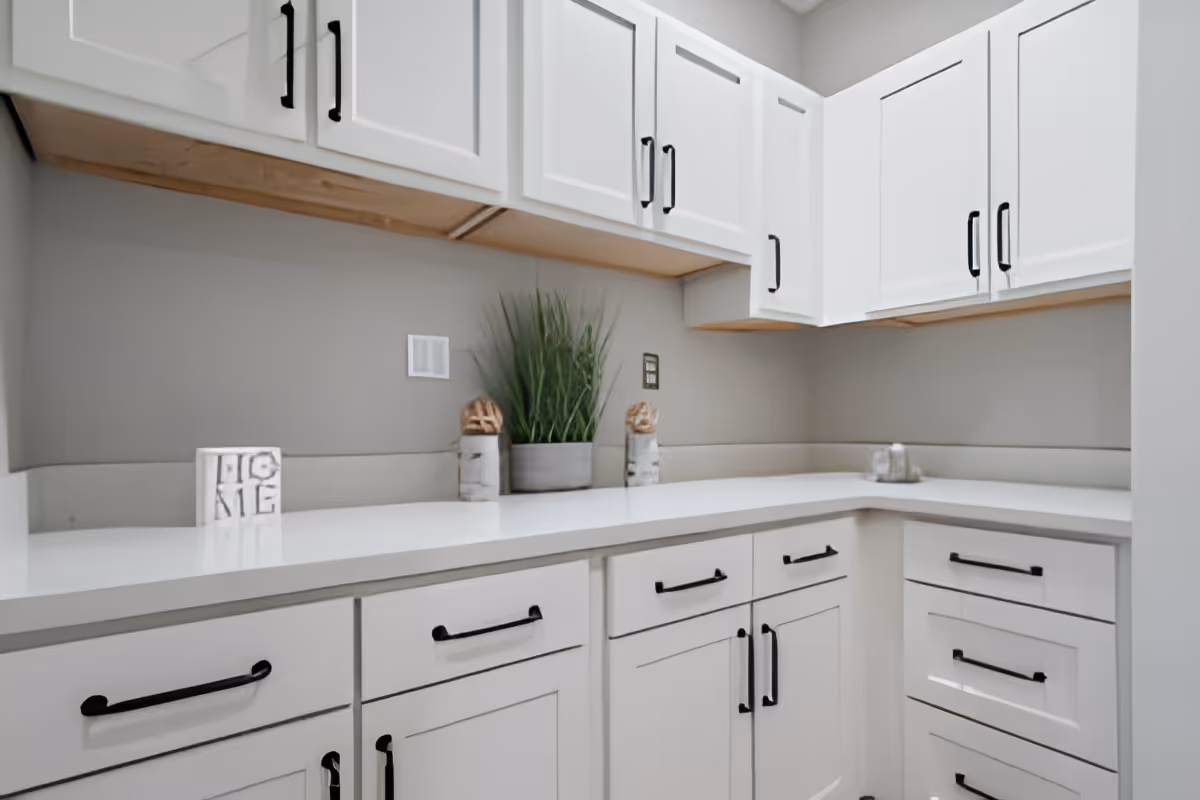 A clean, modern kitchen corner with white cabinets and drawers featuring black handles. The countertop is white and decorated with a small potted plant and two decorative wooden pieces. There is also a small block with the word 'HOME' on it.
