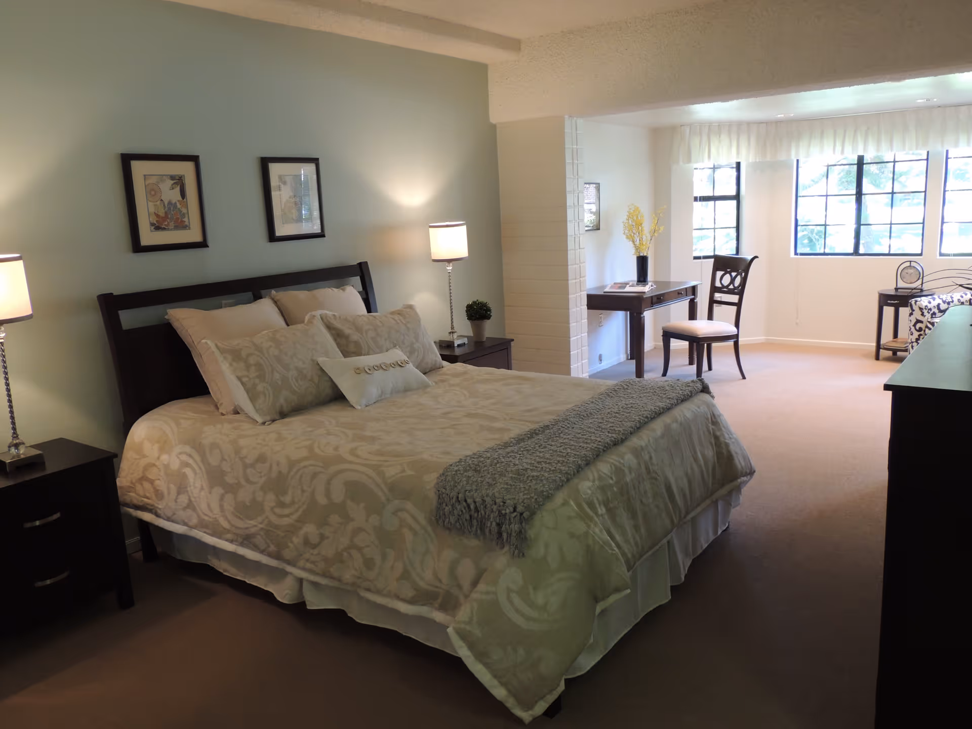 A spacious bedroom with a large bed covered in beige patterned bedding and multiple pillows. There are two bedside tables with lamps on either side of the bed. The room has light-colored walls, two framed pictures above the bed, and a sitting area with a desk, chair, and a small table near large windows letting in natural light.