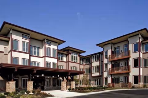 Three-story senior living facility with balconies and a covered entrance, blue sky overhead.