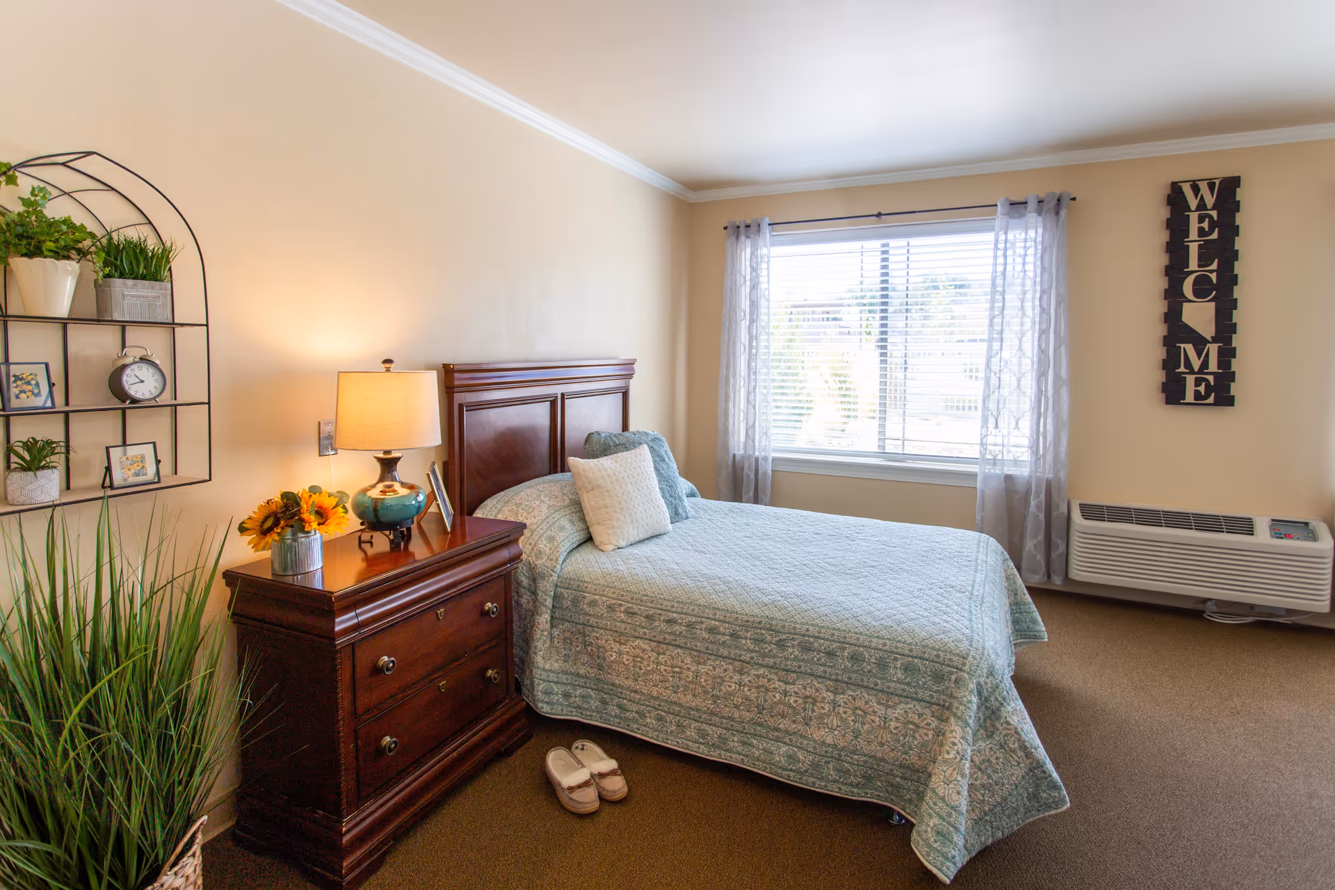 A cozy bedroom with a single bed covered in a light blue patterned quilt and two pillows. Next to the bed is a wooden nightstand with a lamp, a small vase of sunflowers, and a picture frame. On the wall above the nightstand is a decorative shelf holding small plants, a clock, and framed photos. A large window with sheer curtains lets in natural light. On the wall near the window is a vertical sign that reads 'WELCOME'. A pair of slippers is placed on the floor beside the bed.