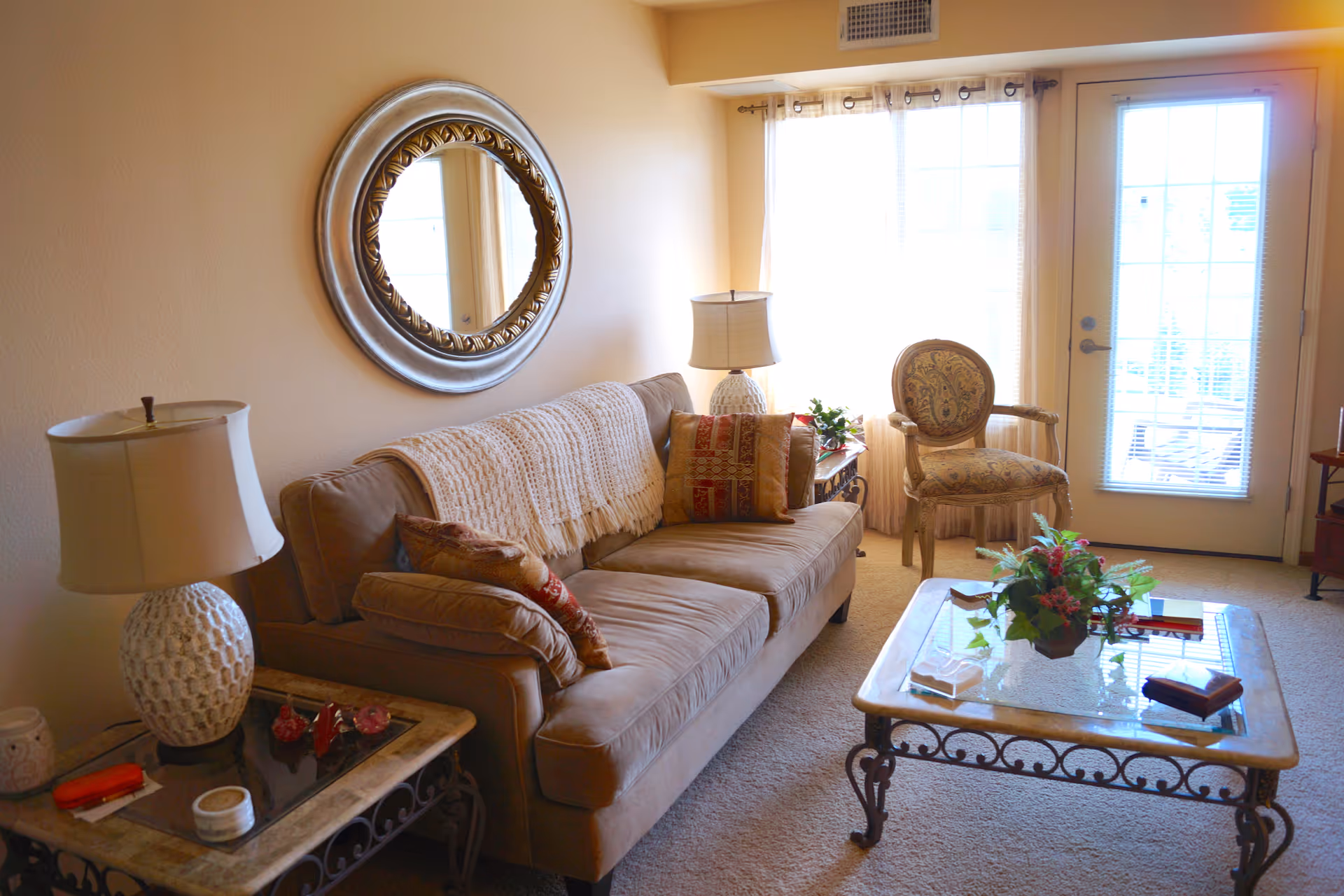 Sunlit living room with a beige sofa, decorative pillows and throw, a glass-top coffee table with a plant, an upholstered armchair, table lamps, and a round wall mirror near a glass door.