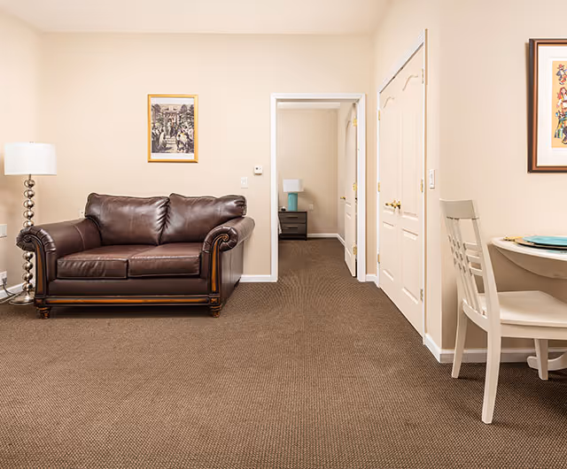 A furnished living area with a brown leather sofa, floor lamp, and small dining table, with a doorway leading to another room.