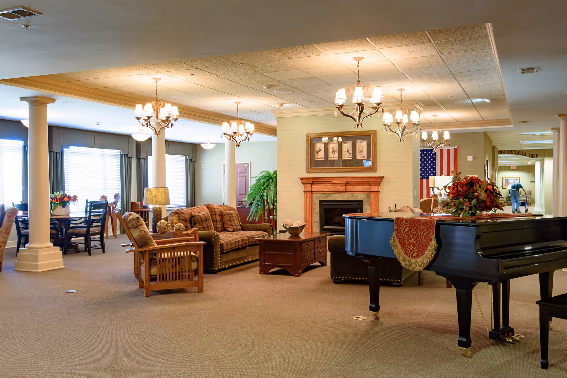 A spacious and well-lit senior living facility common area featuring comfortable seating with plaid sofas and wooden armchairs, a grand piano with a decorative cloth and flower arrangement, a fireplace with framed artwork above it, chandeliers hanging from the ceiling, and large windows with curtains allowing natural light. In the background, an American flag is displayed and a person using a walker is visible near a hallway.