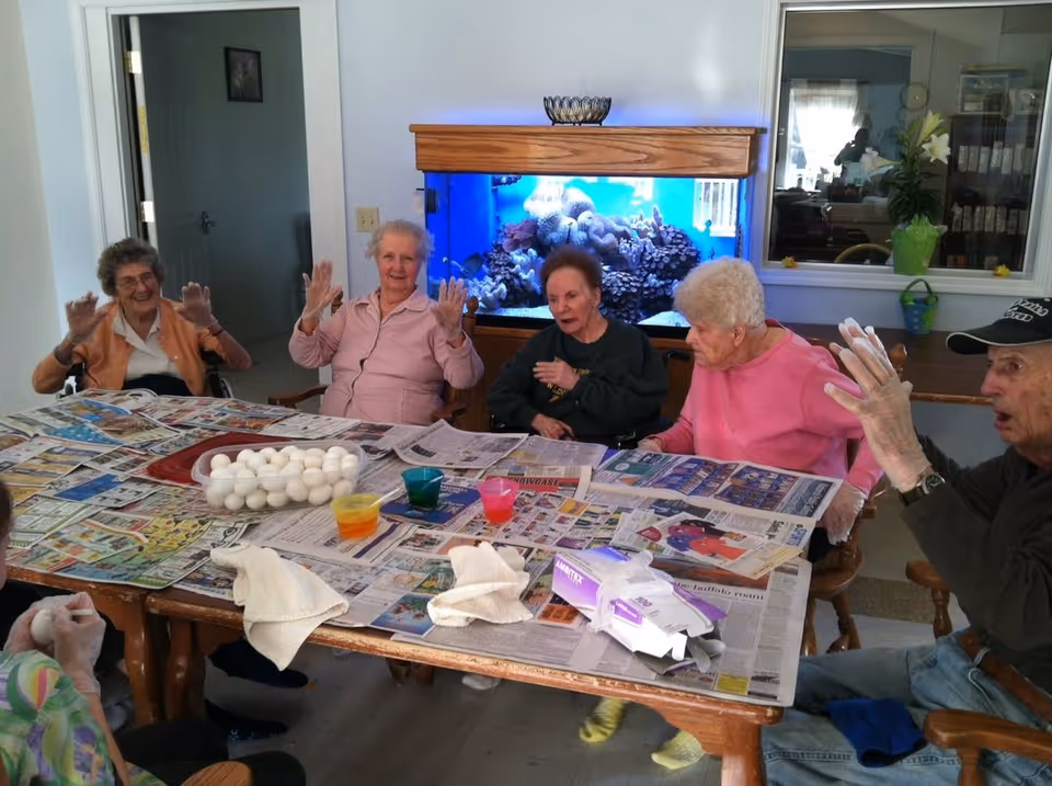 A group of elderly people sitting around a table covered with newspapers, wearing gloves, and preparing to dye eggs. There is a large aquarium with coral and fish in the background, and a window showing another room with shelves and plants.